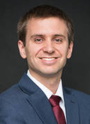 A professional headshot of a person wearing a dark suit, white shirt, and red tie, set against a dark background.