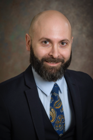 Headshot of a person with a beard, wearing a dark suit, light blue shirt, and patterned tie, set against a textured brown background.