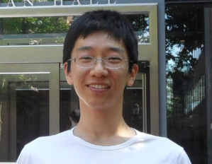 Headshot of a person with glasses, short black hair, and a white t-shirt, smiling in front of a modern building entrance.