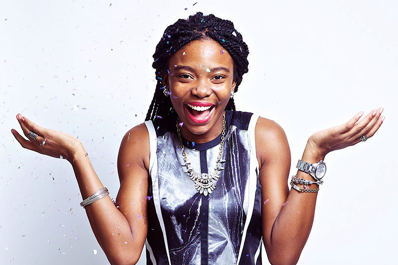 A person joyfully poses with arms outstretched and confetti falling around them, wearing a sleeveless top and accessorized with bracelets and a necklace.