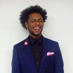 A person with curly hair is smiling while wearing a dark blue suit, a black shirt, and pink accessories, including a pocket square and boutonnière.