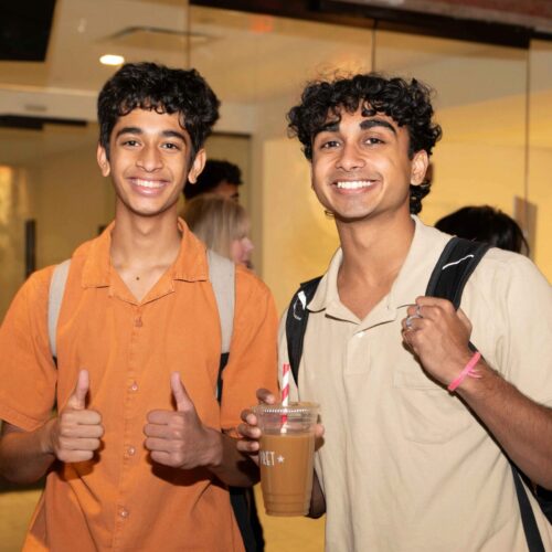 Two smiling individuals with backpacks, one giving thumbs up and the other holding a drink, standing indoors with people in the background.