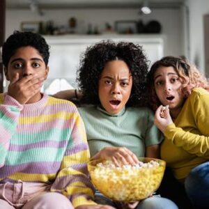 Three people sitting on a couch holding a bowl of popcorn, looking surprised or shocked, possibly watching a movie or TV show.