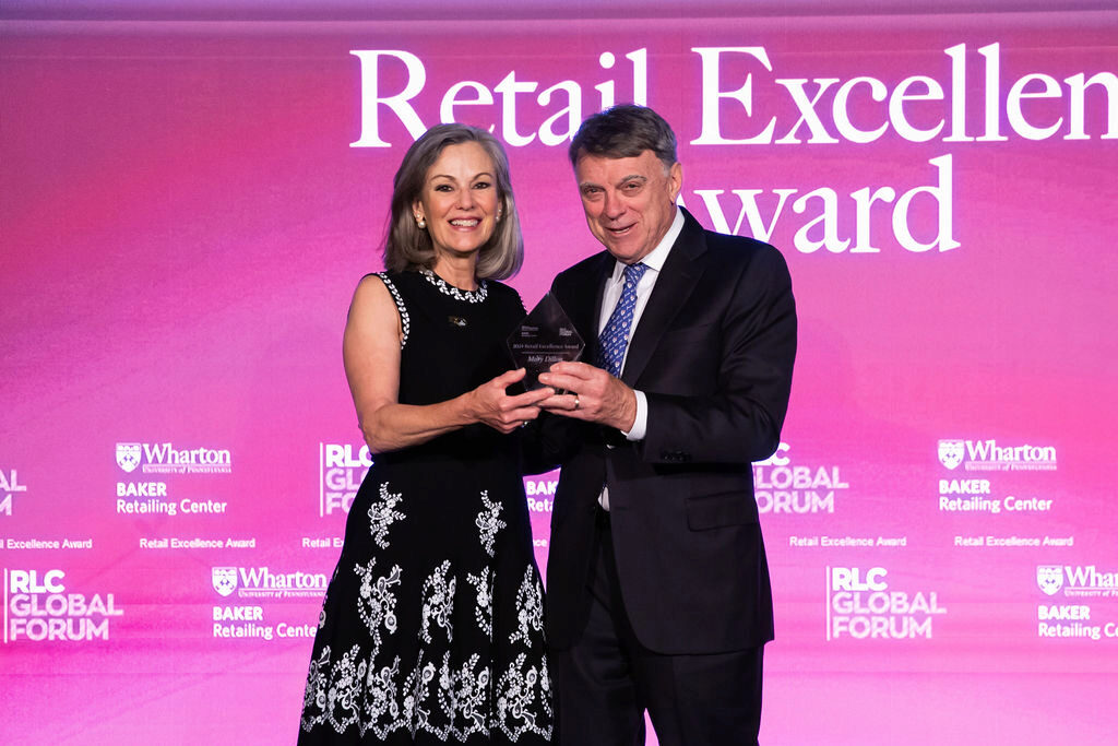 A person in formal attire presents an award to another person against a purple backdrop that says "Retail Excellence Award" and includes branding for Wharton and Baker Retailing Center.