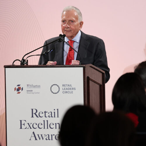 A person speaking at a podium during the Retail Excellence Awards event. The background displays logos for the Wharton Baker Retailing Center and Retail Leaders Circle.