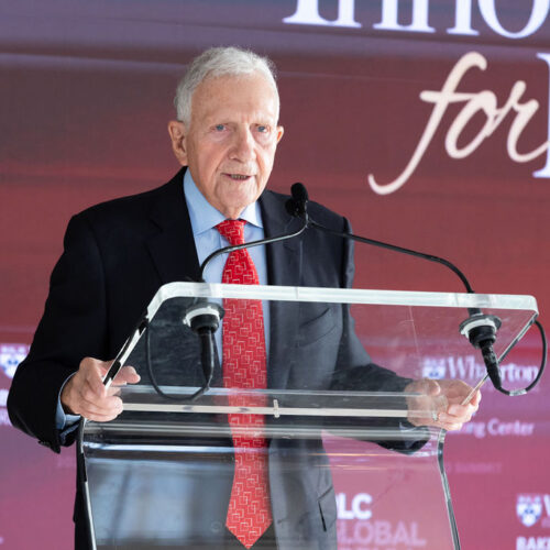 An older individual in a suit and red tie speaks at a clear podium with a microphone. The backdrop has text related to a global forum or summit event.