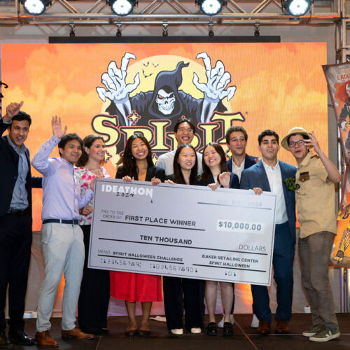 A group of people celebrating on stage, holding a large novelty check for $10,000, labeled as the first-place winner of the Spirit Halloween Challenge. The backdrop features Halloween-themed decorations and graphics.