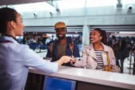 A couple at an airport check-in counter interacting with an airline employee who is handing them documents. The scene is lively with other travelers in the background.