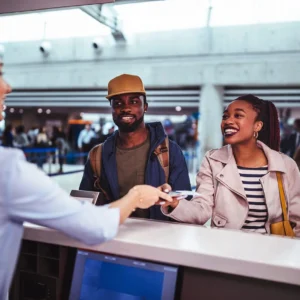 A couple at an airport check-in counter interacting with an airline employee who is handing them documents. The scene is lively with other travelers in the background.