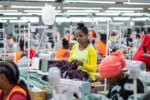 A large garment factory with several individuals working at sewing machines. One person in the foreground is focusing on fabric, while others are working at stations in the background.