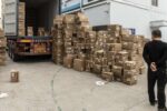 A person stands near a large pile of cardboard boxes, some stacked and others scattered, next to a shipping container on a trailer.