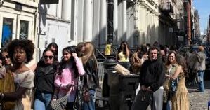 A diverse group of people stand in line outside on a city street, suggesting a busy urban setting or event.
