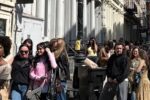 A long line of people standing on a sidewalk in an urban area, possibly waiting for an event or store opening, with buildings in the background.