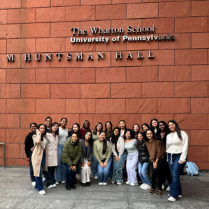 A group of people, posing in front of a building with "The Wharton School University of Pennsylvania JM Huntsman Hall" sign on the wall. They appear to be smiling and standing together.