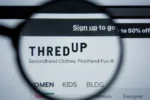 Website screenshot featuring the ThredUp logo under a magnifying glass, with the slogan "Secondhand Clothes. Firsthand Fun." visible. Options for sections like "Women," "Kids," and "Blog" are shown.