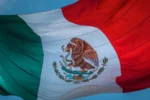 The image shows the flag of Mexico, featuring vertical stripes in green, white, and red with a coat of arms depicting an eagle holding a snake on a cactus in the center stripe.