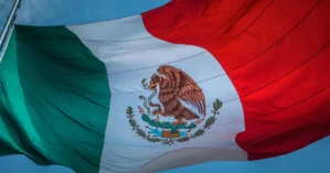 The image shows the flag of Mexico, featuring vertical stripes in green, white, and red with a coat of arms depicting an eagle holding a snake on a cactus in the center stripe.