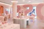 A modern, brightly lit cosmetics store with minimalistic white and pink decor. Displays feature various beauty products, mirrors, and a central floral arrangement. Large pink numbers and a logo are on the walls.