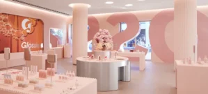 A modern, brightly lit cosmetics store with minimalistic white and pink decor. Displays feature various beauty products, mirrors, and a central floral arrangement. Large pink numbers and a logo are on the walls.