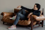 A person is sitting casually on a brown leather sofa, wearing a dark blazer, jeans, and colorful sneakers, appearing relaxed and smiling.