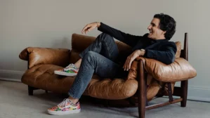 A person is sitting casually on a brown leather sofa, wearing a dark blazer, jeans, and colorful sneakers, appearing relaxed and smiling.
