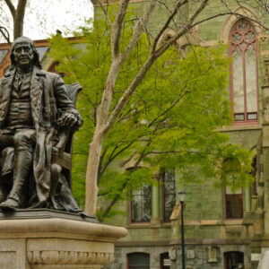 A bronze statue of a person sitting on a chair, located in front of a historic building with arched windows and ornate architecture. Trees with budding leaves are nearby.