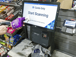 A person scanning an item at a self-checkout kiosk in a store, with a "Start Scanning" prompt displayed and shopping bags on the counter.