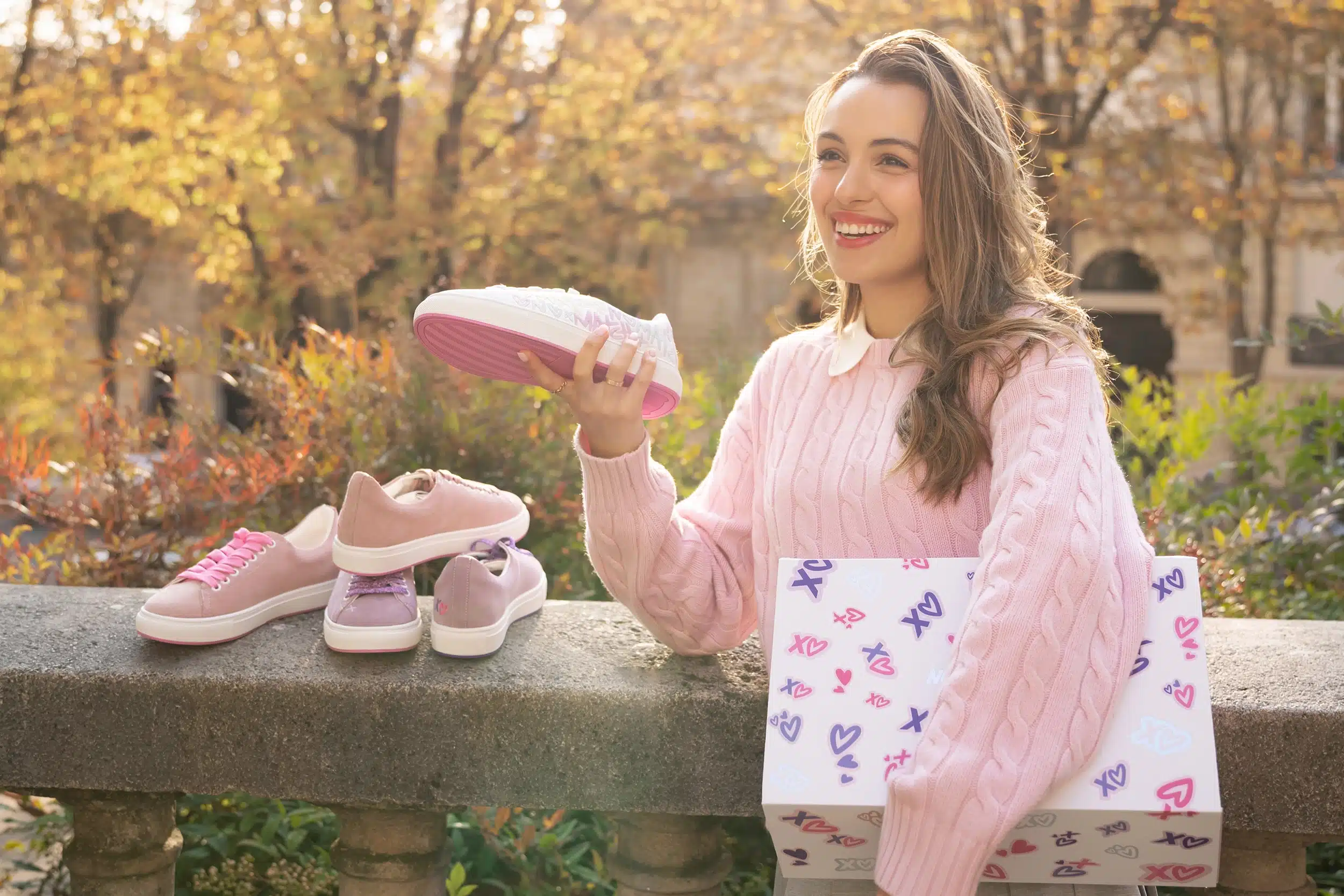 A person in a pink sweater is joyfully holding a shoe and a shoebox with an assortment of shoes placed on a stone ledge. Autumn foliage is in the background.