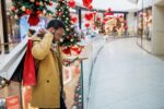 Person in a tan coat holding shopping bags and looking at a phone in a festive mall with holiday decorations and Christmas trees.