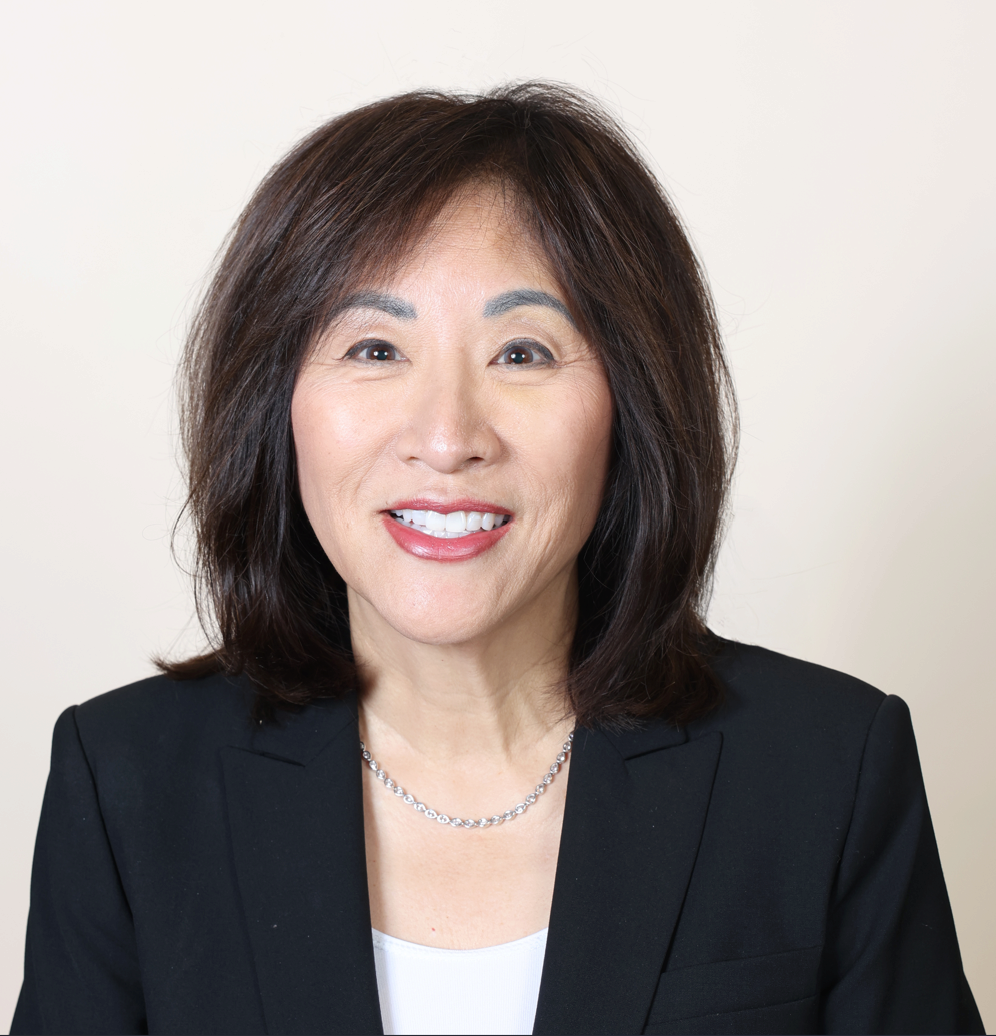 Screenshot 2026-01-22 135907 Headshot of person with shoulder-length dark hair, wearing a black blazer, white top, and a necklace, against a plain background.