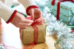 Person gift-wrapping presents with festive paper and ribbon, surrounded by holiday decorations like tinsel on a wooden table.